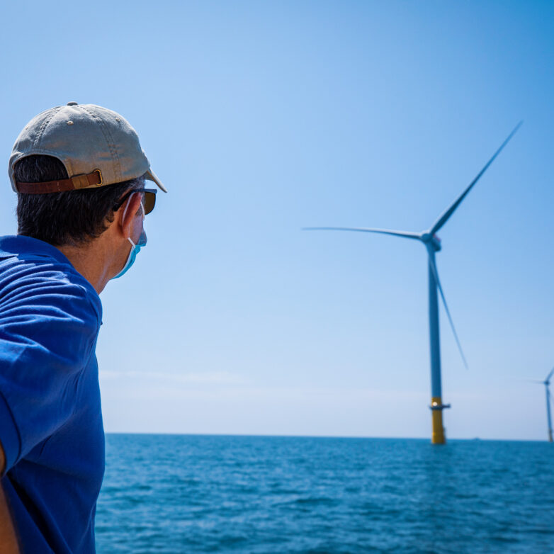 Man with mask and hat on looks out into body of water at wind farm
