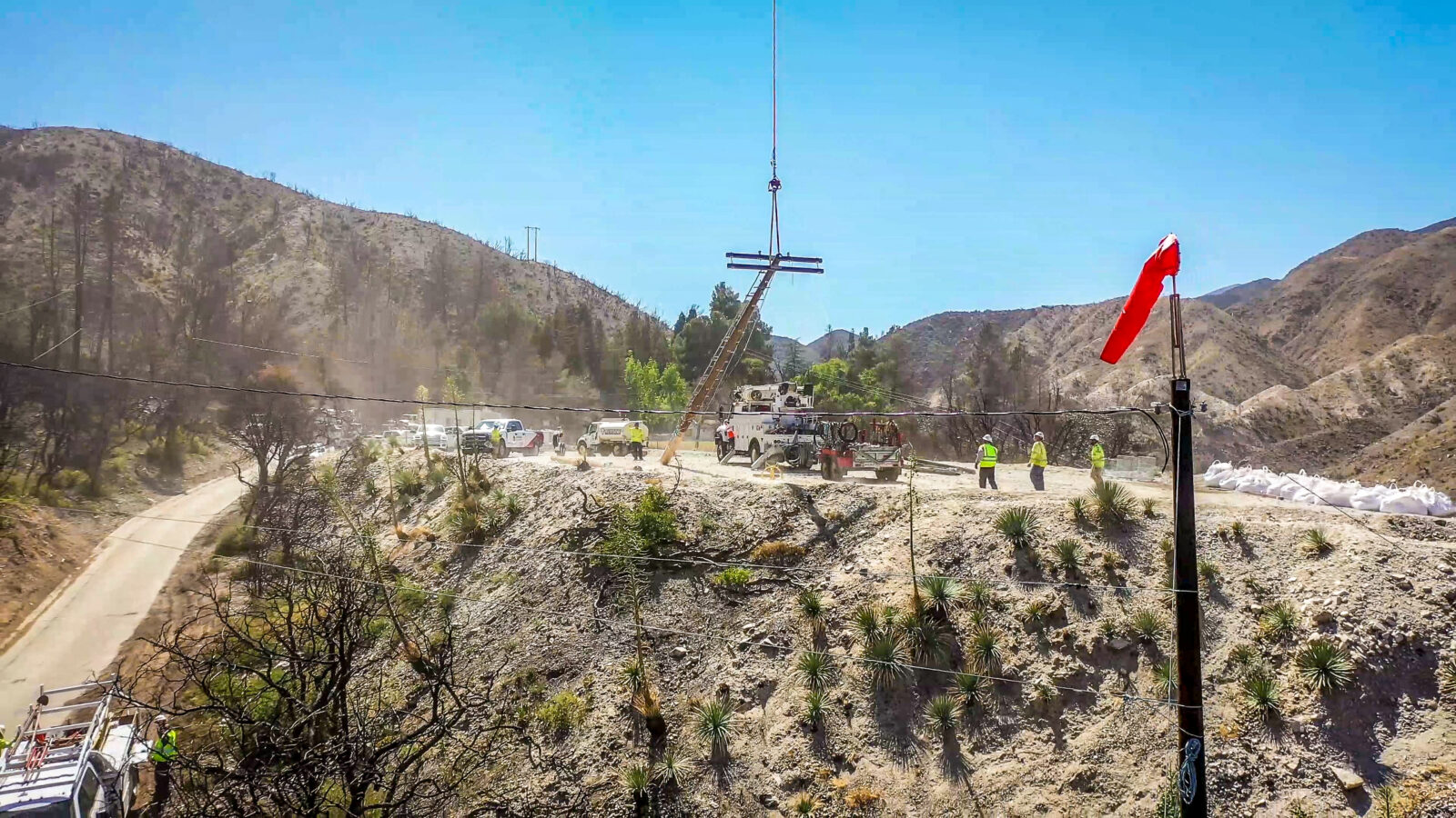 Trucks and electrical workers on a ridge as a power line pole is raised by a helicopter.