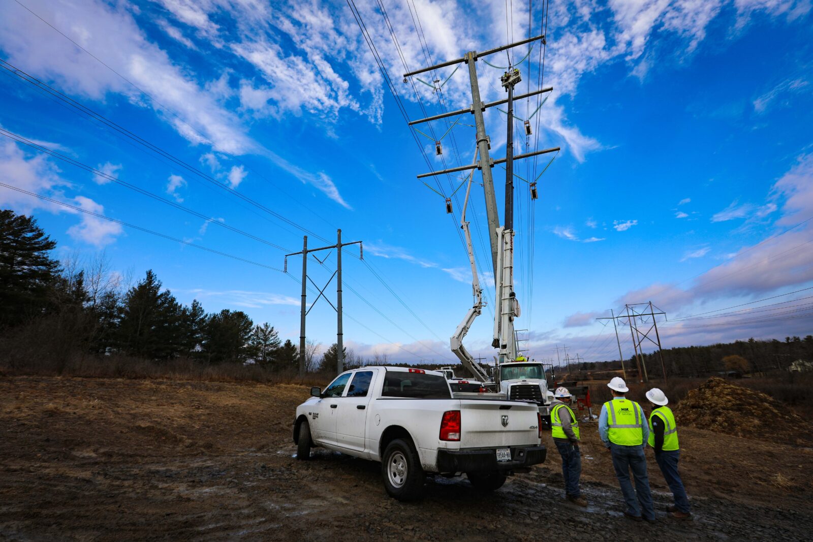 Three men in hard hats stand underneath a tranmission line and near a white pickup truck.