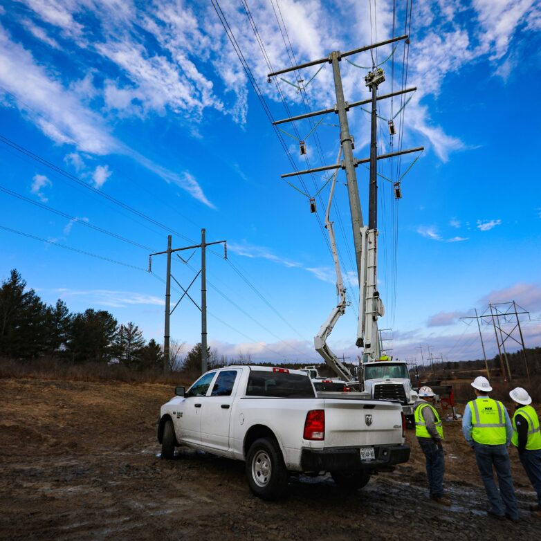 Three men in hard hats stand underneath a tranmission line and near a white pickup truck.