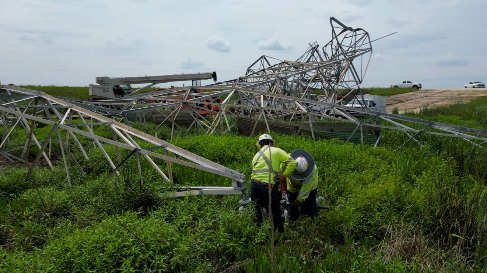 A storm damaged transmission tower lies on the ground. Two construction workers examine a portion of the steel.